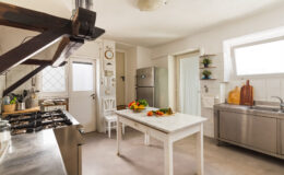 Bright and modern kitchen featuring a marble-topped table with fresh vegetables, stainless steel appliances, and wooden accents. The space includes a gas stove, a large refrigerator, and ample natural light from windows, creating an inviting atmosphere for cooking and dining.