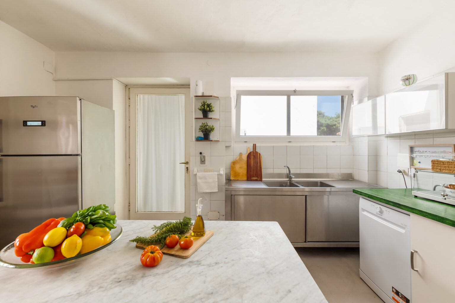 Modern kitchen featuring stainless steel appliances, a green countertop, and a bowl of fresh fruits and vegetables on a marble island, with ample natural light from a window. | Only on SopranoVillas