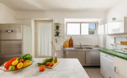Modern kitchen featuring stainless steel appliances, a green countertop, and a bowl of fresh fruits and vegetables on a marble island, with ample natural light from a window.