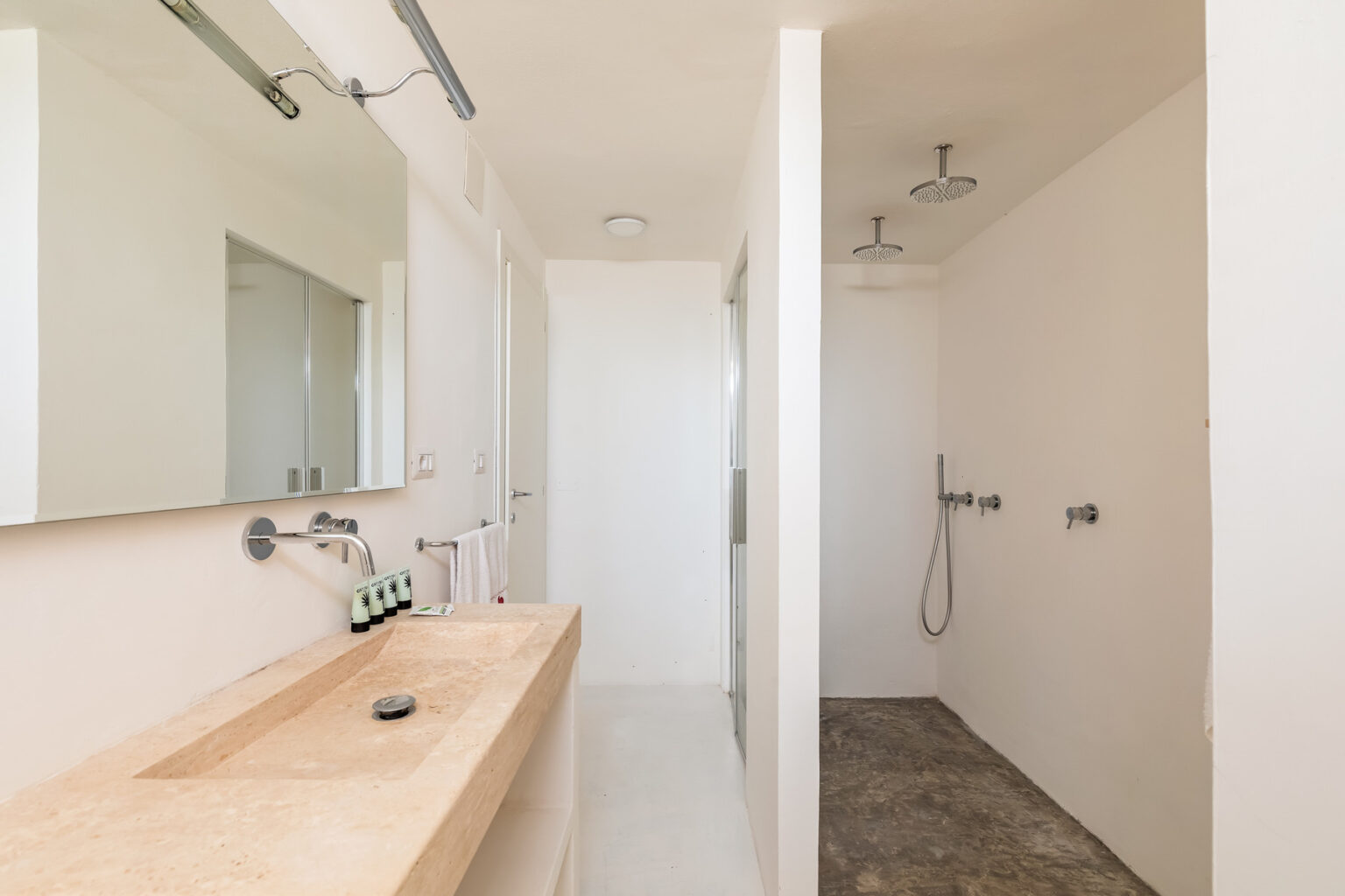 Modern bathroom interior featuring a minimalist design with a stone sink, large mirror, and dual showerheads. The space includes a walk-in shower, light-colored walls, and a neutral flooring that enhances the contemporary aesthetic. | Only on SopranoVillas