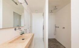 Modern bathroom interior featuring a minimalist design with a stone sink, large mirror, and dual showerheads. The space includes a walk-in shower, light-colored walls, and a neutral flooring that enhances the contemporary aesthetic.