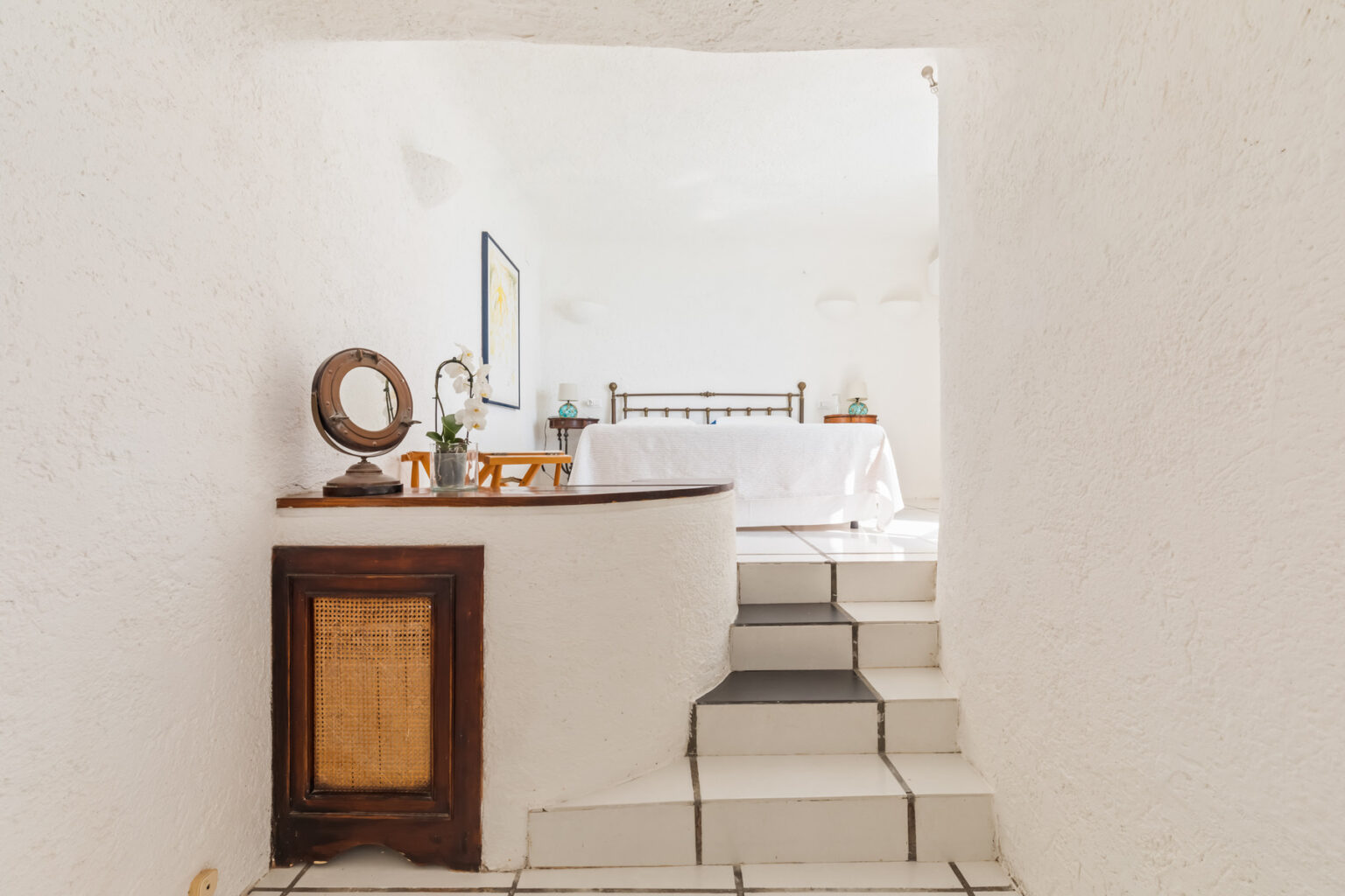 Interior view of a bright, minimalist bedroom featuring white walls and tiled flooring, with a wooden console table displaying a mirror and a vase of orchids, and a staircase leading to the sleeping area. | Only on SopranoVillas