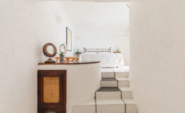 Interior view of a bright, minimalist bedroom featuring white walls and tiled flooring, with a wooden console table displaying a mirror and a vase of orchids, and a staircase leading to the sleeping area.