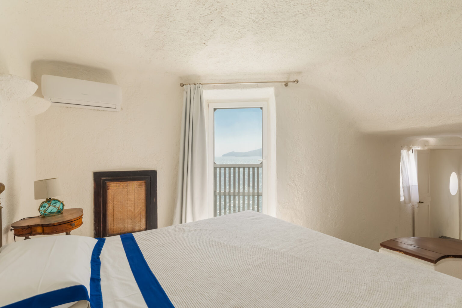 Cozy bedroom interior featuring a bed with white linens and a blue accent, an air conditioning unit on the wall, and a window offering a scenic view of the sea and mountains. Natural light illuminates the room, enhancing the serene atmosphere. | Only on SopranoVillas