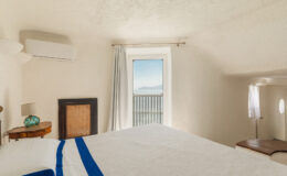 Cozy bedroom interior featuring a bed with white linens and a blue accent, an air conditioning unit on the wall, and a window offering a scenic view of the sea and mountains. Natural light illuminates the room, enhancing the serene atmosphere.