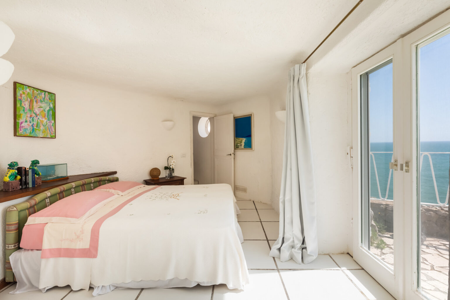 Bright and airy bedroom with a white color scheme, featuring a cozy double bed with pink accents, a small wooden nightstand, and artwork on the walls. Large windows provide a stunning ocean view, enhancing the serene atmosphere of the space. | Only on SopranoVillas