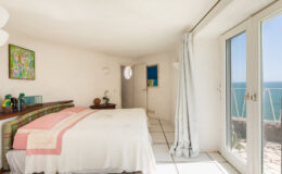 Bright and airy bedroom with a white color scheme, featuring a cozy double bed with pink accents, a small wooden nightstand, and artwork on the walls. Large windows provide a stunning ocean view, enhancing the serene atmosphere of the space.