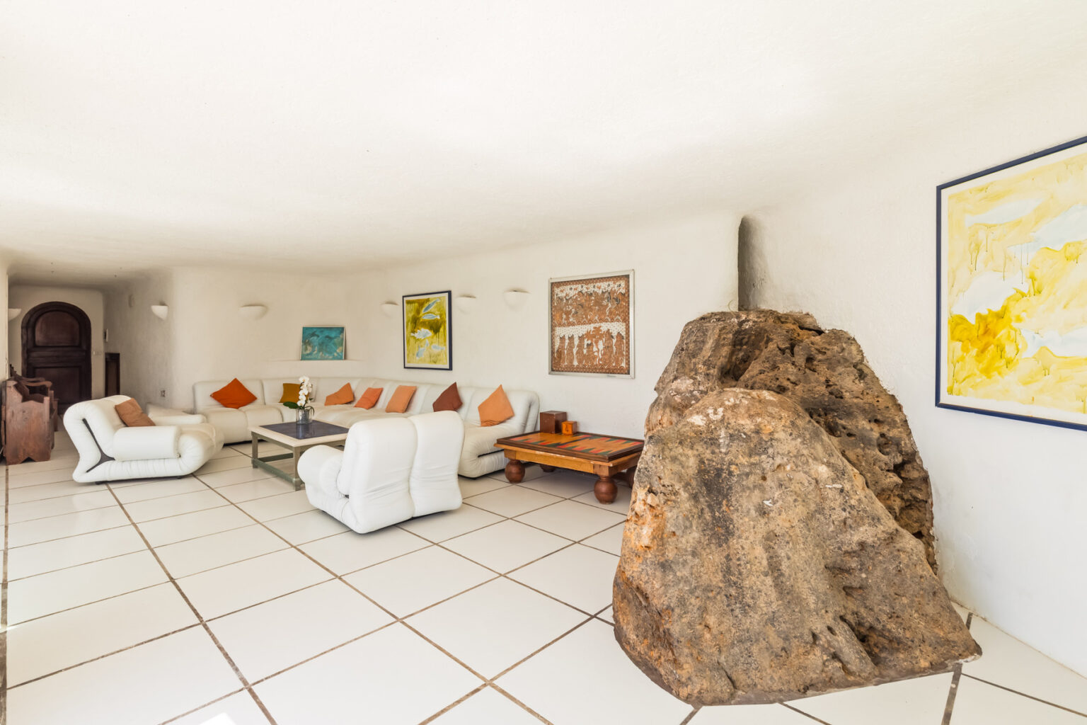 Modern living room featuring minimalist white furniture, colorful artwork, and a large natural rock as a focal point, enhancing the contemporary design. Bright, spacious layout with tiled flooring and warm accents. | Only on SopranoVillas