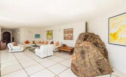 Modern living room featuring minimalist white furniture, colorful artwork, and a large natural rock as a focal point, enhancing the contemporary design. Bright, spacious layout with tiled flooring and warm accents.