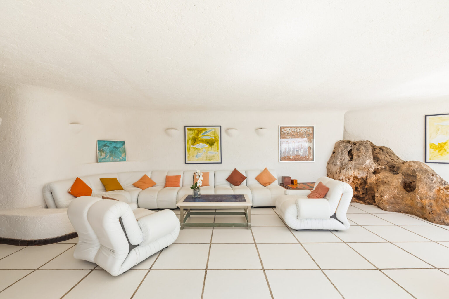 Modern living room with a white color scheme, featuring a curved white sectional sofa adorned with colorful cushions, a glass coffee table, and artwork on the walls, complemented by a natural rock formation. | Only on SopranoVillas