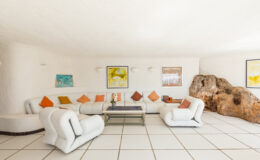 Modern living room with a white color scheme, featuring a curved white sectional sofa adorned with colorful cushions, a glass coffee table, and artwork on the walls, complemented by a natural rock formation.