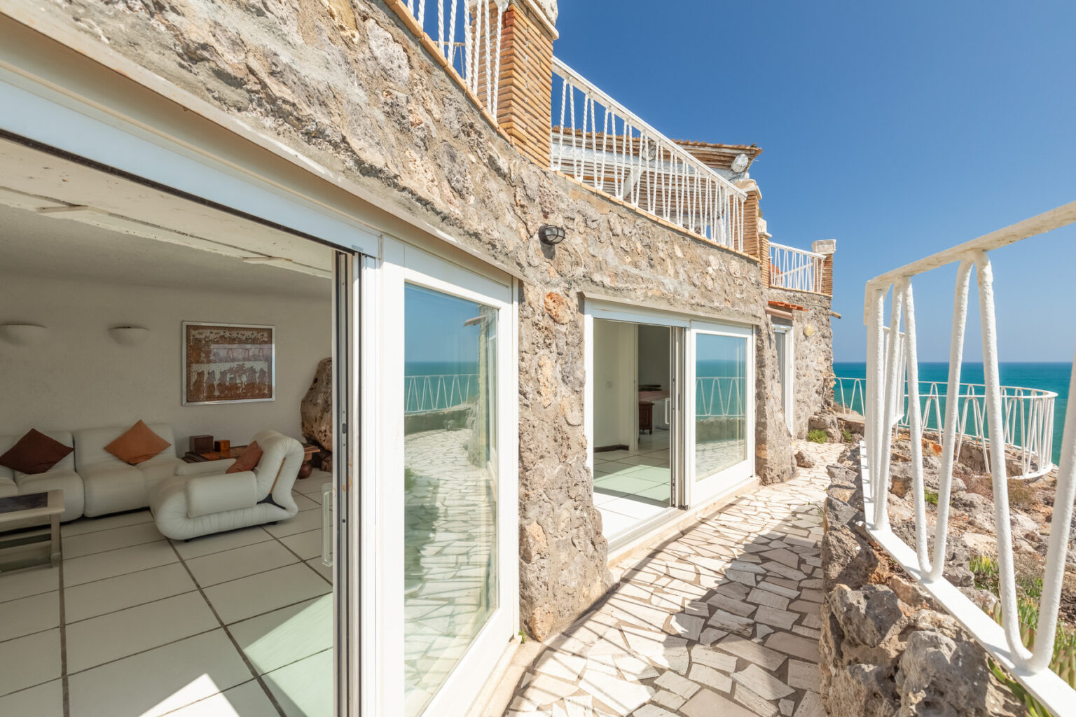 Coastal stone villa with large glass doors, offering scenic ocean views and a sunlit terrace. The image showcases the modern interior visible through the open doors, framed by a natural stone exterior. | Only on SopranoVillas