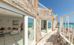Coastal stone villa with large glass doors, offering scenic ocean views and a sunlit terrace. The image showcases the modern interior visible through the open doors, framed by a natural stone exterior.
