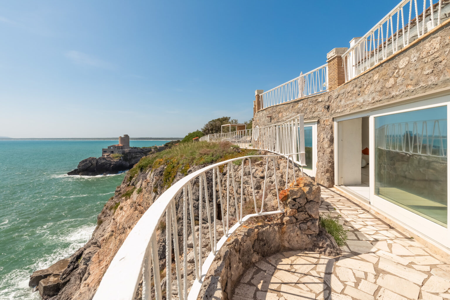 Coastal view from a modern stone balcony overlooking the ocean, with clear blue skies and a nearby rocky shoreline. | Only on SopranoVillas