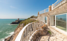 Coastal view from a modern stone balcony overlooking the ocean, with clear blue skies and a nearby rocky shoreline.