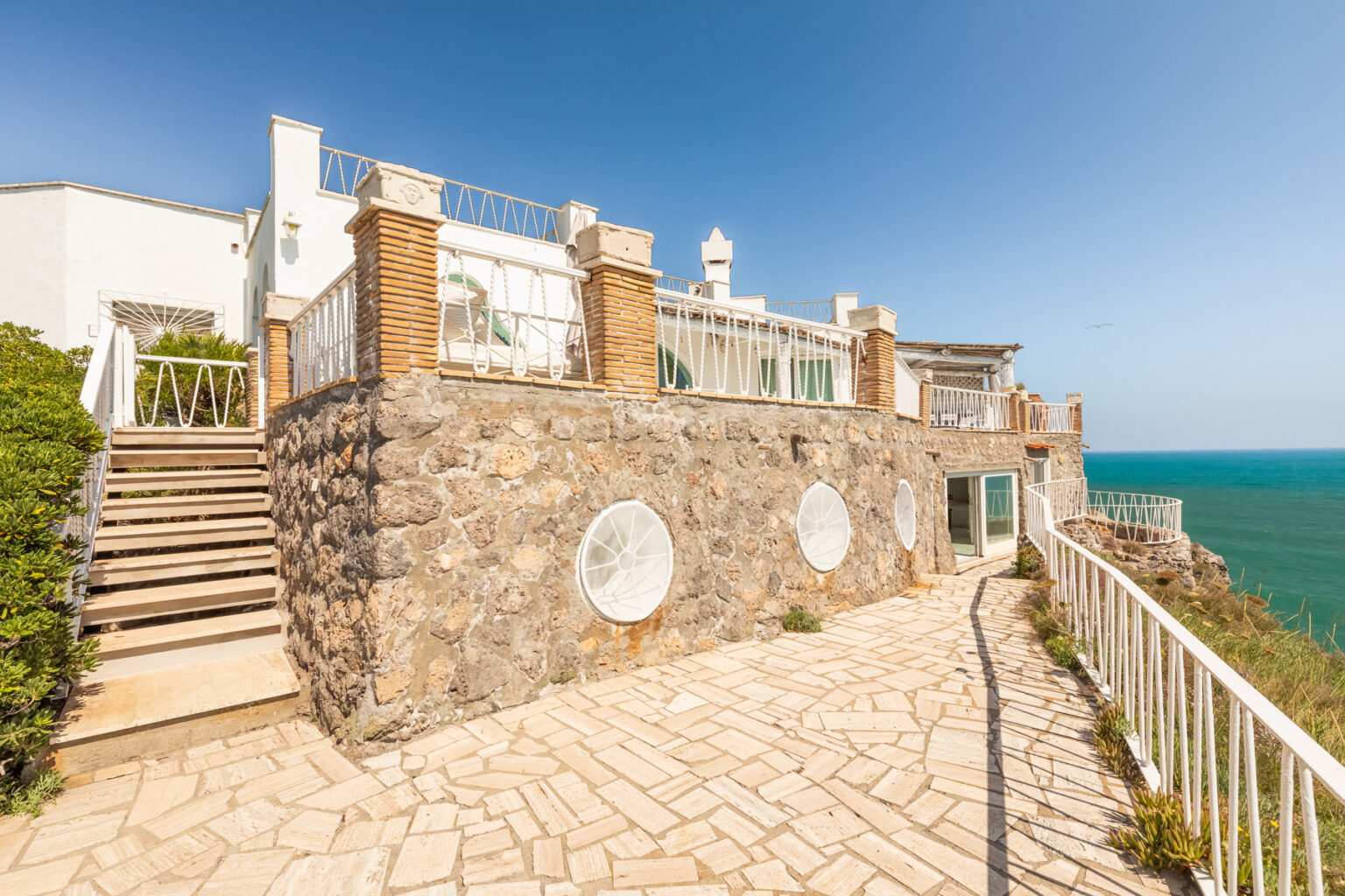 Stunning coastal villa with stone walls and a spacious terrace overlooking the ocean, featuring white railings and steps leading to the entrance. Bright blue sky enhances the scenic view. | Only on SopranoVillas