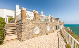 Stunning coastal villa with stone walls and a spacious terrace overlooking the ocean, featuring white railings and steps leading to the entrance. Bright blue sky enhances the scenic view.