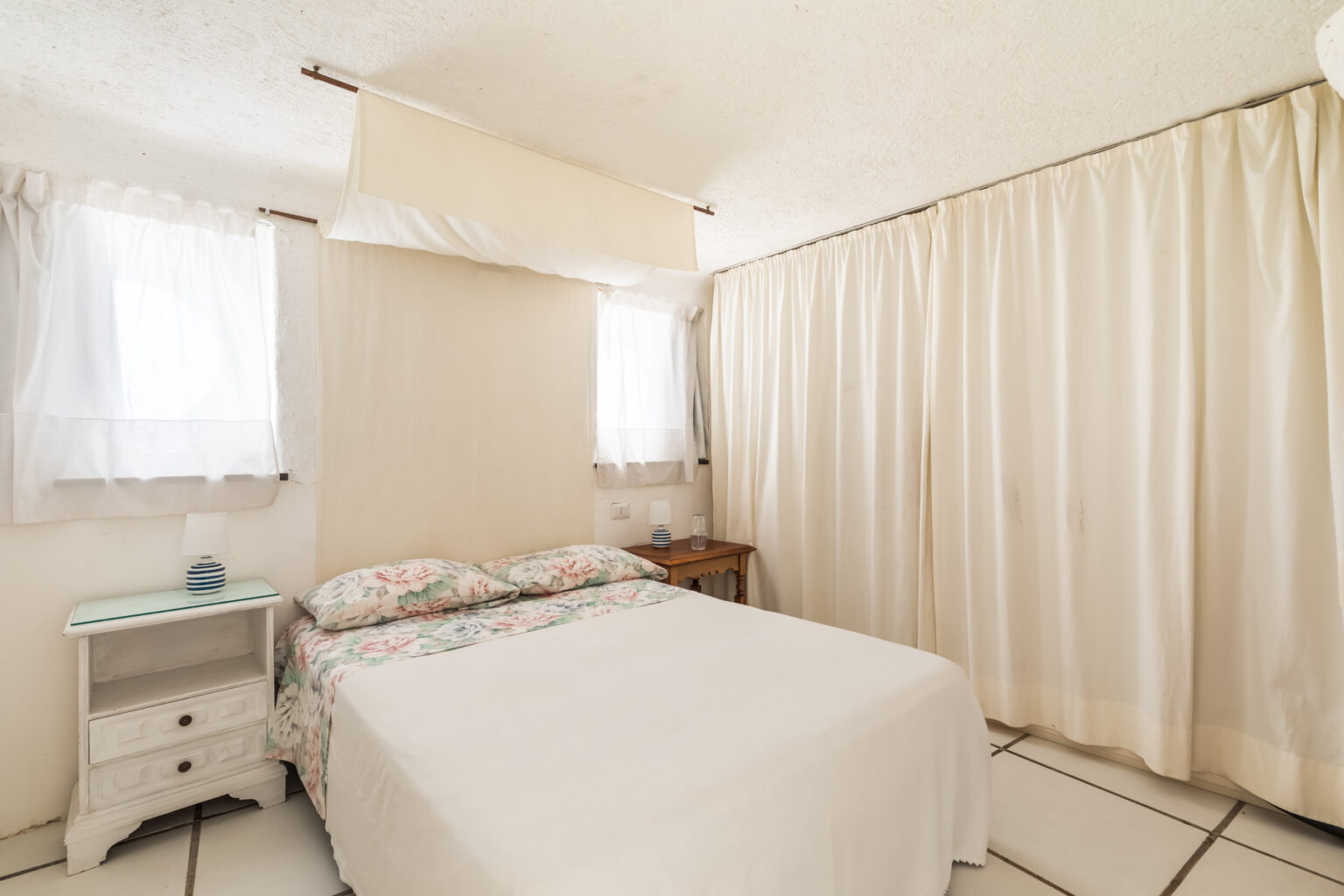 Cozy bedroom featuring a double bed with floral bedding, a bedside table, and natural light streaming through sheer curtains. The room has a neutral color palette and simple decor, creating a relaxing atmosphere. | Only on SopranoVillas