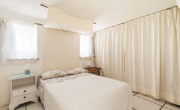 Cozy bedroom featuring a double bed with floral bedding, a bedside table, and natural light streaming through sheer curtains. The room has a neutral color palette and simple decor, creating a relaxing atmosphere.