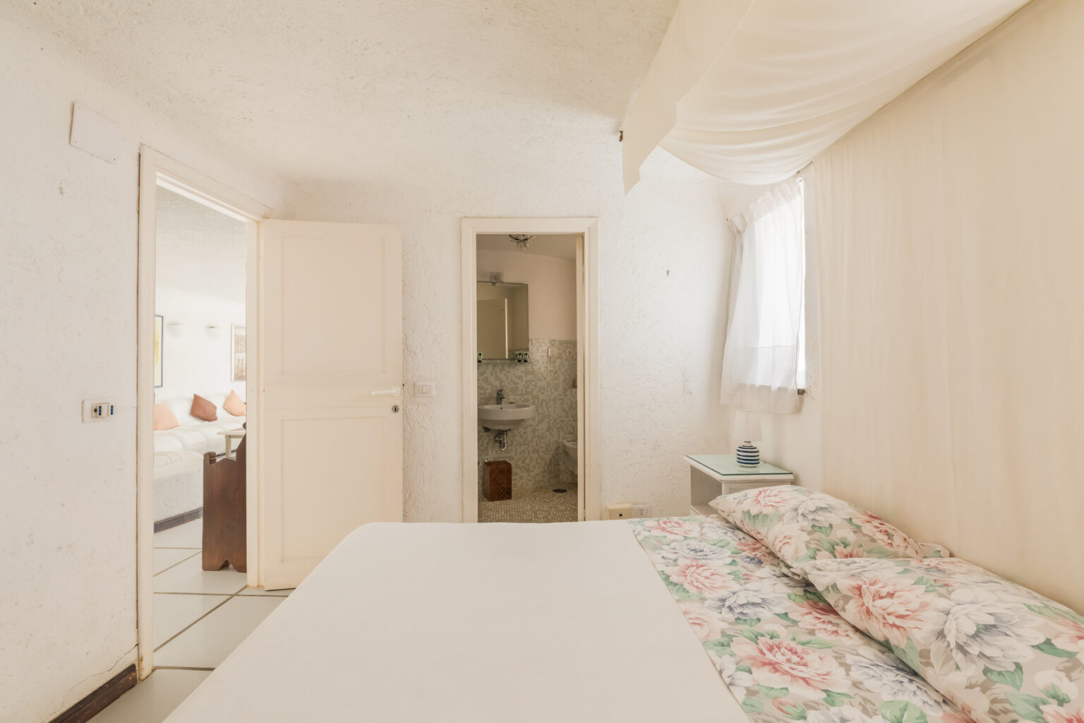 Cozy bedroom interior featuring a bed with floral bedding, white walls, and natural light streaming through a window, with a bathroom visible through an open door. | Only on SopranoVillas