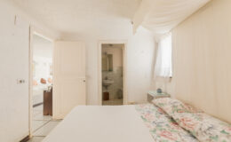 Cozy bedroom interior featuring a bed with floral bedding, white walls, and natural light streaming through a window, with a bathroom visible through an open door.