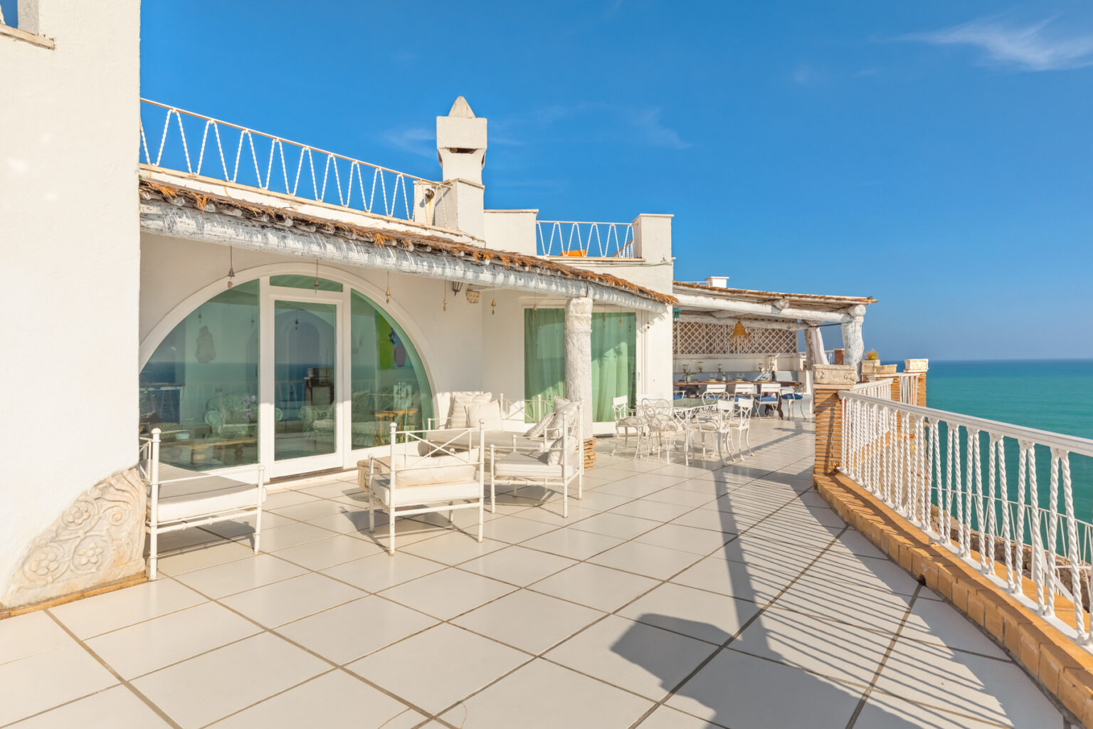 Spacious seaside terrace with white furniture and panoramic ocean views, featuring glass doors leading to an airy interior, ideal for relaxation and outdoor dining. | Only on SopranoVillas