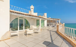 Spacious seaside terrace with white furniture and panoramic ocean views, featuring glass doors leading to an airy interior, ideal for relaxation and outdoor dining.
