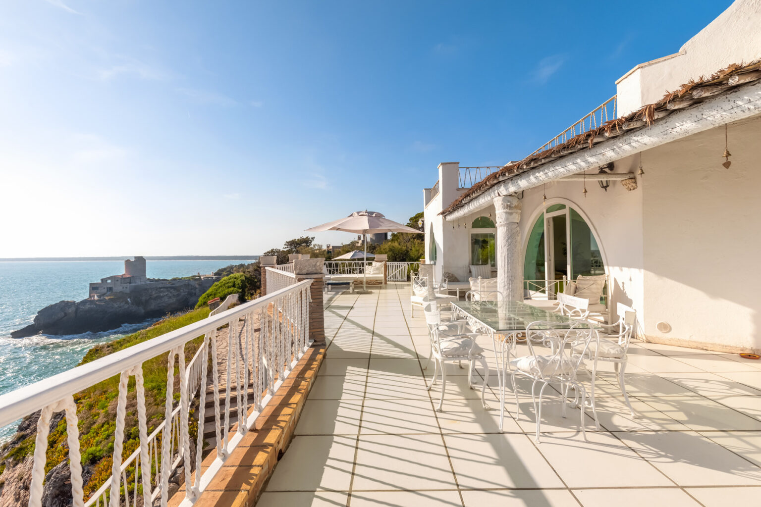 Spacious oceanfront terrace with white wrought iron furniture, offering stunning views of the sea and nearby rocky coastline under a clear blue sky. | Only on SopranoVillas
