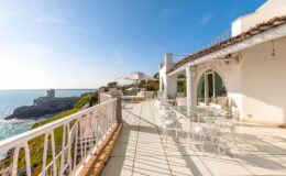 Spacious oceanfront terrace with white wrought iron furniture, offering stunning views of the sea and nearby rocky coastline under a clear blue sky.