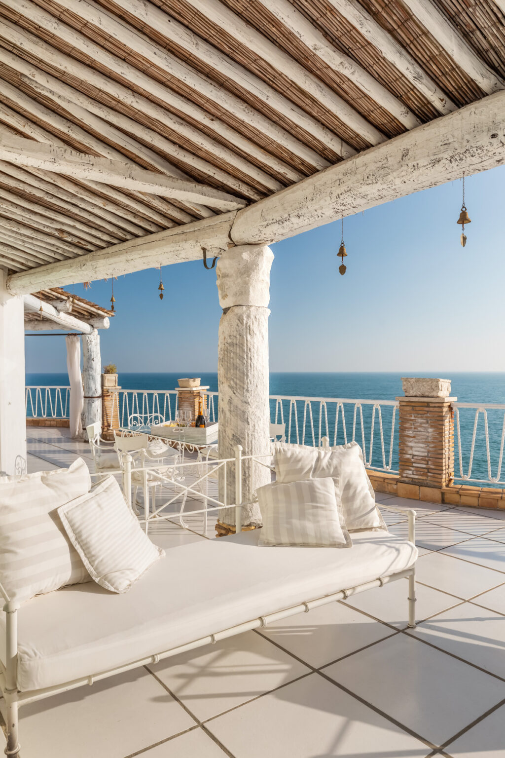 Cozy outdoor terrace with a white metal lounge chair and pillows, featuring a stunning view of the ocean and a clear blue sky. The space is adorned with rustic wooden beams and decorative bells, creating a relaxing atmosphere ideal for enjoying seaside moments. | Only on SopranoVillas