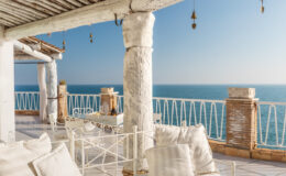 Cozy outdoor terrace with a white metal lounge chair and pillows, featuring a stunning view of the ocean and a clear blue sky. The space is adorned with rustic wooden beams and decorative bells, creating a relaxing atmosphere ideal for enjoying seaside moments.