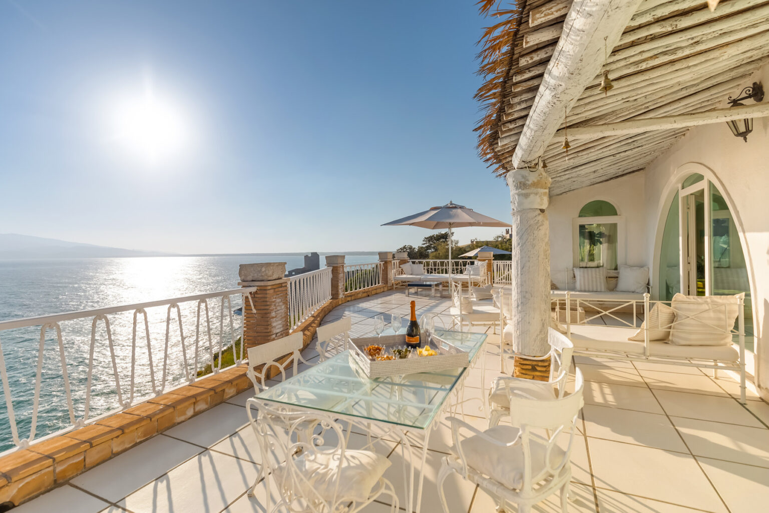 Luxury oceanfront terrace with glass table, white seating, and a bottle of champagne, overlooking sparkling waters and a clear blue sky. Perfect for relaxing and enjoying stunning sunset views. | Only on SopranoVillas