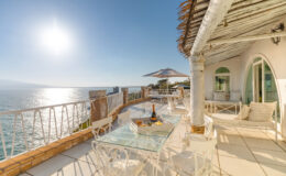 Luxury oceanfront terrace with glass table, white seating, and a bottle of champagne, overlooking sparkling waters and a clear blue sky. Perfect for relaxing and enjoying stunning sunset views.