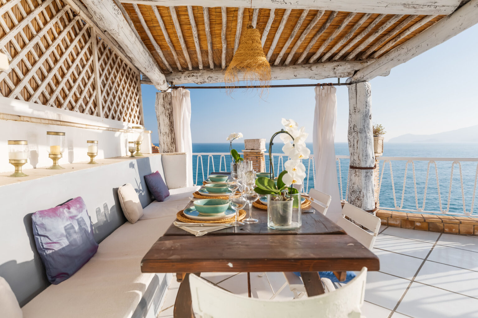 Cozy outdoor dining area with a wooden table set for a meal, featuring elegant tableware and decorative flowers, overlooking a stunning ocean view under a rustic pergola. | Only on SopranoVillas