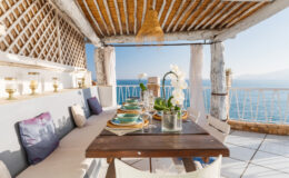 Cozy outdoor dining area with a wooden table set for a meal, featuring elegant tableware and decorative flowers, overlooking a stunning ocean view under a rustic pergola.