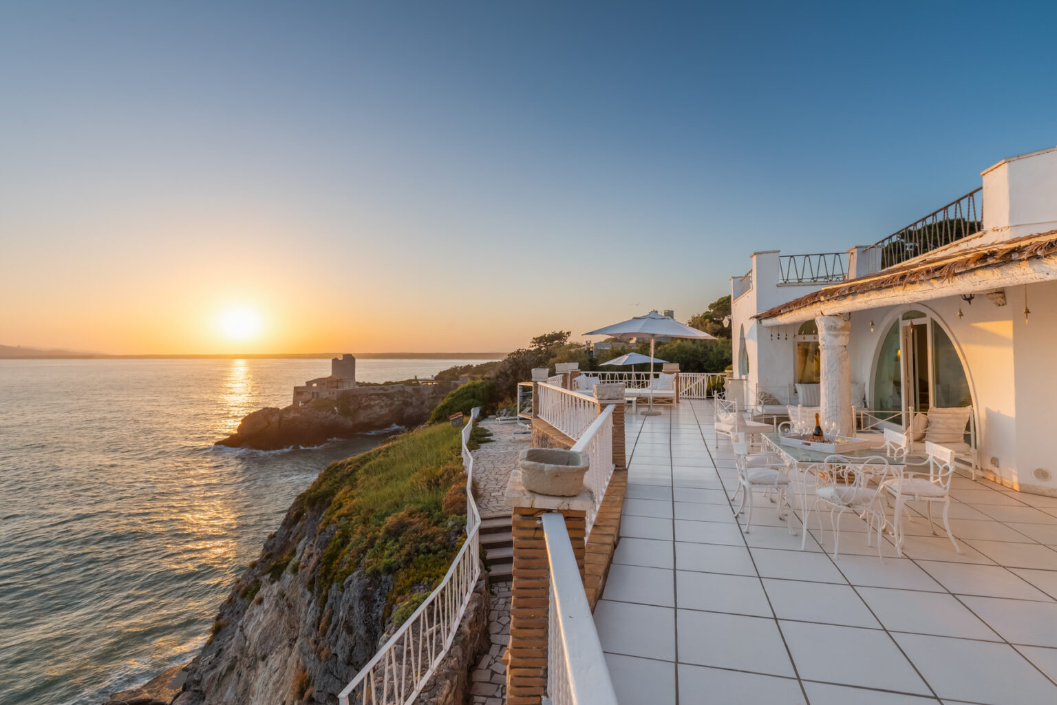 Scenic oceanfront terrace at sunset, featuring modern white architecture, outdoor seating, and panoramic views of the coastline and tranquil waters. | Only on SopranoVillas