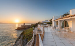 Scenic oceanfront terrace at sunset, featuring modern white architecture, outdoor seating, and panoramic views of the coastline and tranquil waters.