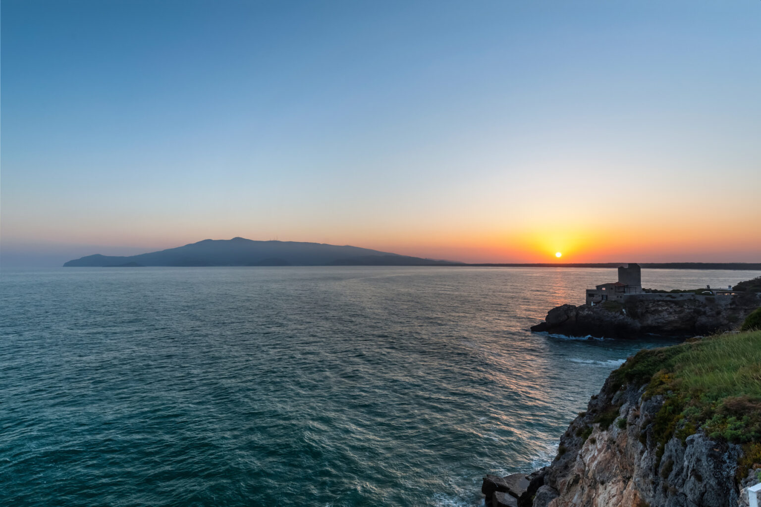 Sunset over the ocean with a view of a distant island, rocky coastline, and calm waters, creating a serene coastal landscape. | Only on SopranoVillas