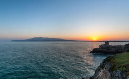 Sunset over the ocean with a view of a distant island, rocky coastline, and calm waters, creating a serene coastal landscape.
