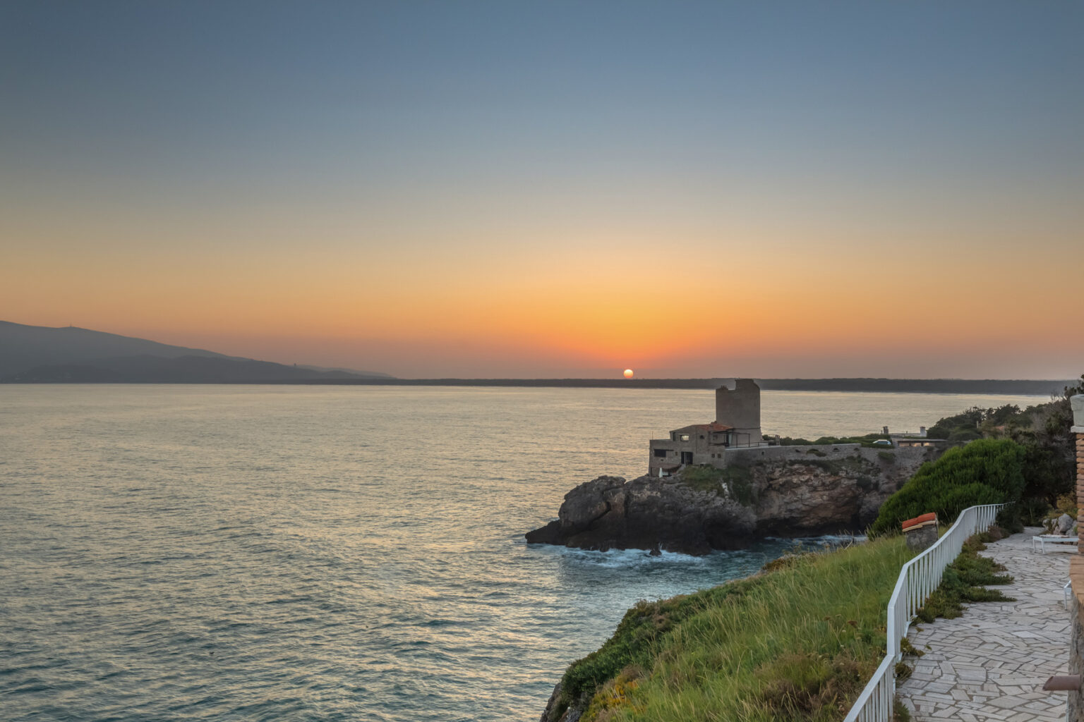Scenic sunset over the ocean with a rocky coastline and a historic tower, highlighting natural beauty and tranquility. | Only on SopranoVillas