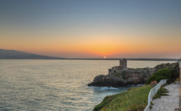 Scenic sunset over the ocean with a rocky coastline and a historic tower, highlighting natural beauty and tranquility.