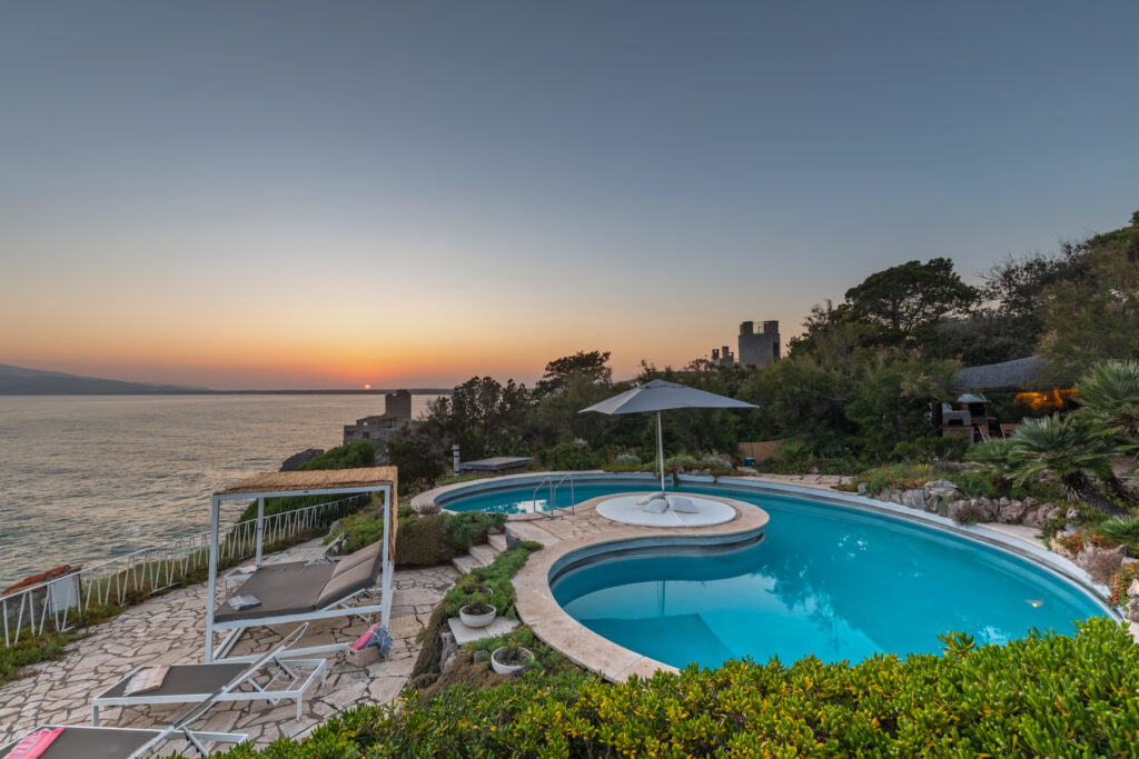 Luxury villa with a stunning infinity pool overlooking the ocean at sunset, surrounded by lush greenery and comfortable lounge chairs. | Only on SopranoVillas
