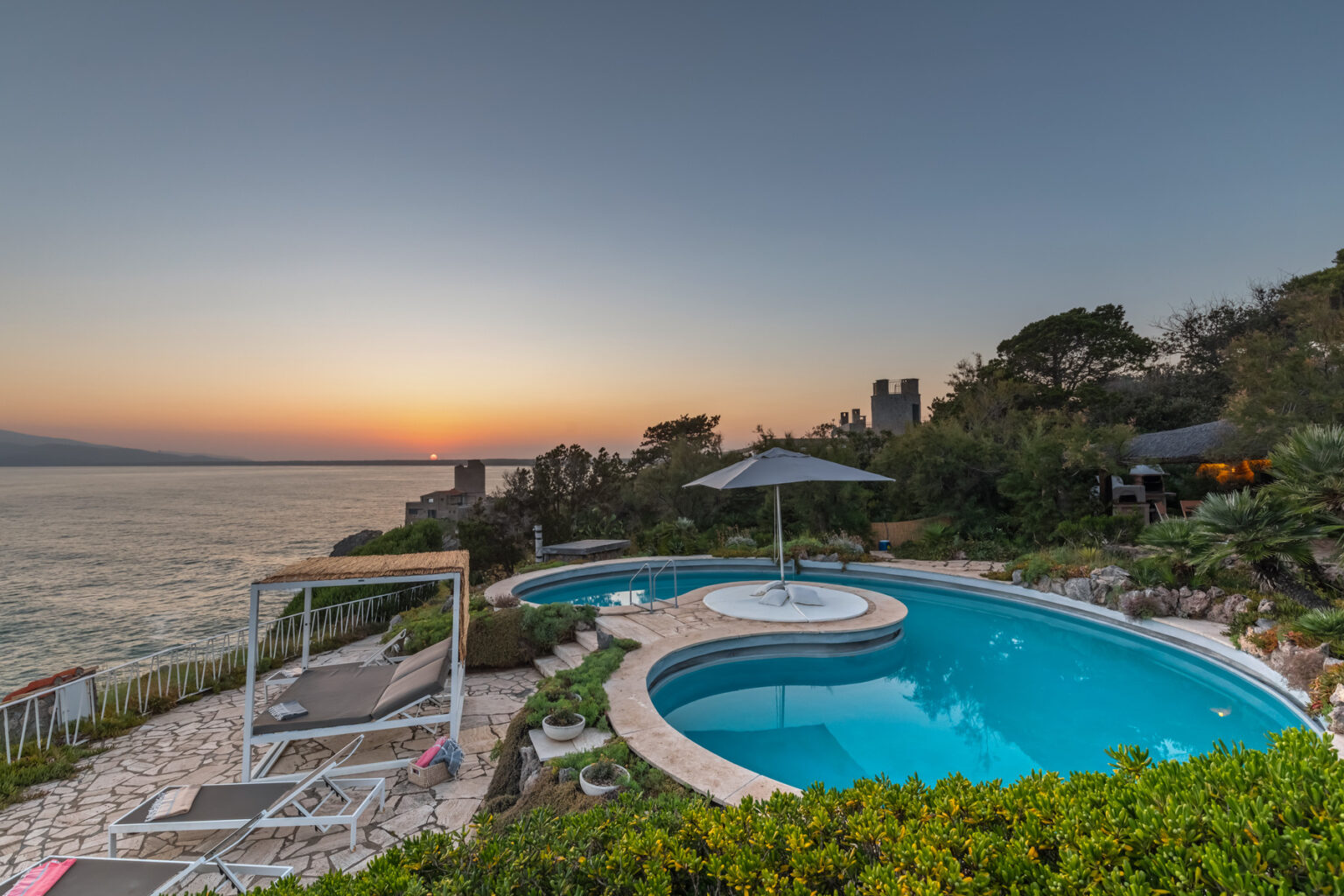 Luxury villa with a stunning infinity pool overlooking the ocean at sunset, surrounded by lush greenery and comfortable lounge chairs. | Only on SopranoVillas