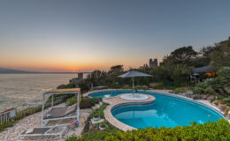Luxury villa with a stunning infinity pool overlooking the ocean at sunset, surrounded by lush greenery and comfortable lounge chairs.