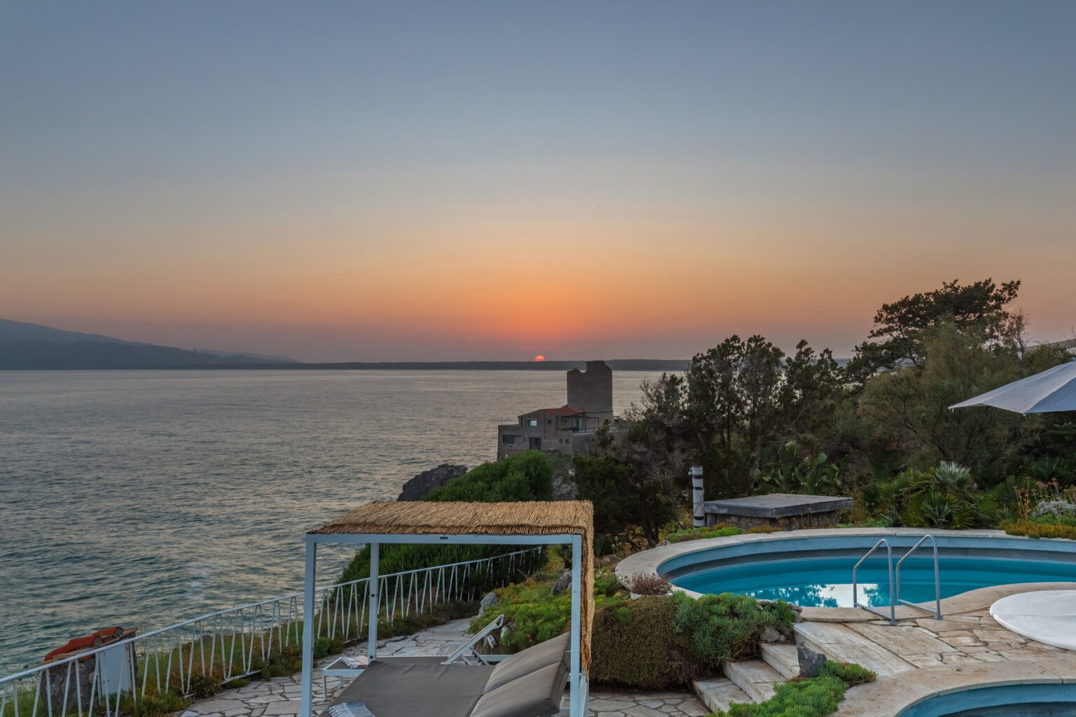 Scenic sunset view over calm waters with a swimming pool in the foreground, surrounded by lush greenery and a distant shoreline. Ideal for promoting vacation rentals or coastal retreats. | Only on SopranoVillas