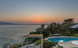 Scenic sunset view over calm waters with a swimming pool in the foreground, surrounded by lush greenery and a distant shoreline. Ideal for promoting vacation rentals or coastal retreats.