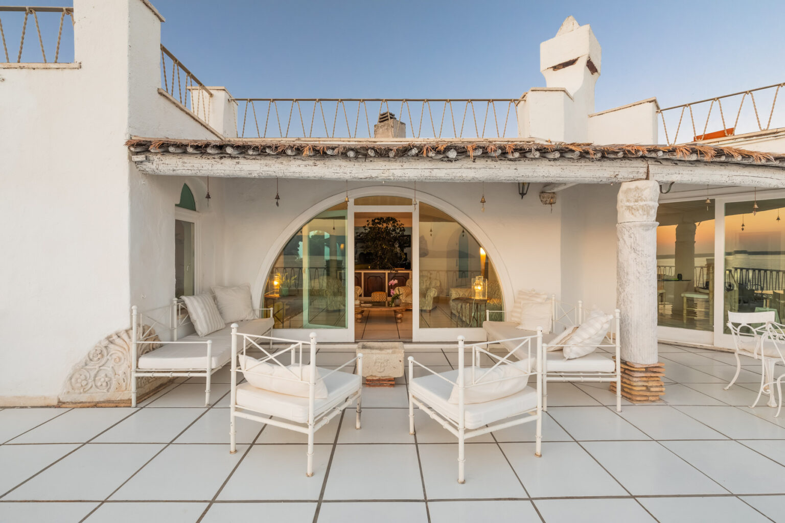 Modern outdoor seating area with white chairs and a curved glass door, showcasing a stylish rooftop terrace of a contemporary villa. The setting features a clean, spacious design with a warm evening ambiance. | Only on SopranoVillas