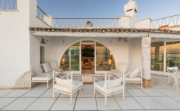 Modern outdoor seating area with white chairs and a curved glass door, showcasing a stylish rooftop terrace of a contemporary villa. The setting features a clean, spacious design with a warm evening ambiance.