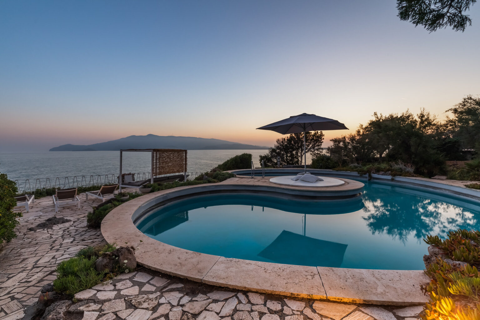 Scenic view of a luxurious poolside at sunset, featuring a circular pool, a relaxing hot tub, and an umbrella, surrounded by lush greenery and overlooking the ocean. | Only on SopranoVillas