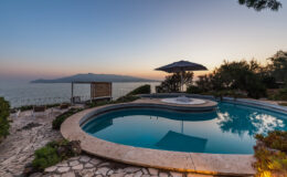 Scenic view of a luxurious poolside at sunset, featuring a circular pool, a relaxing hot tub, and an umbrella, surrounded by lush greenery and overlooking the ocean.